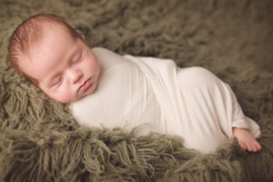 A cute little newborn boy snuggled and wrapped in white while laying on a green flokati rug during his newborn photography session.
